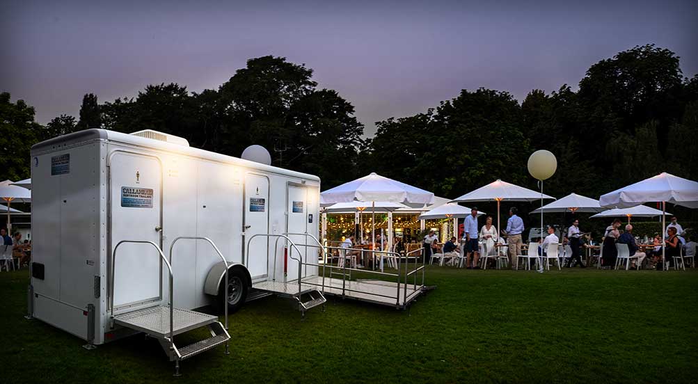 Horizon ADA Restroom Trailer with Outside Lights on, at an Outdoor Venue with People at Various Tented Tables at Dusk